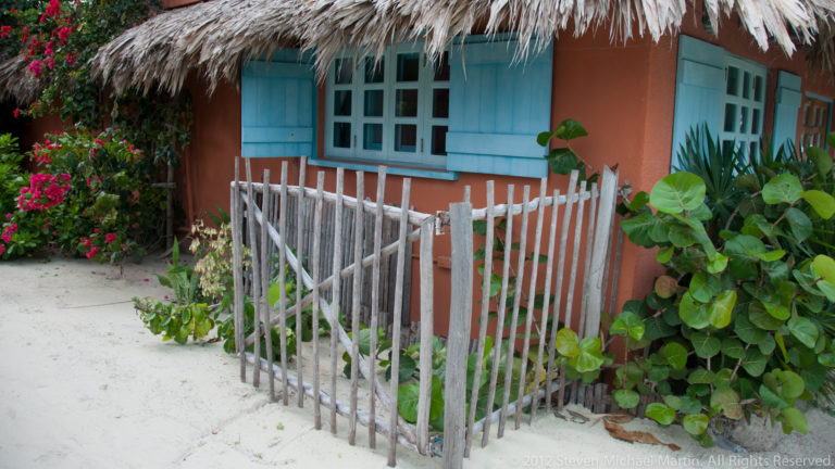 Coral-colored cottage with pale blue windows, thatched roof, rustic picket fence, and tropical plants along a sandy path.