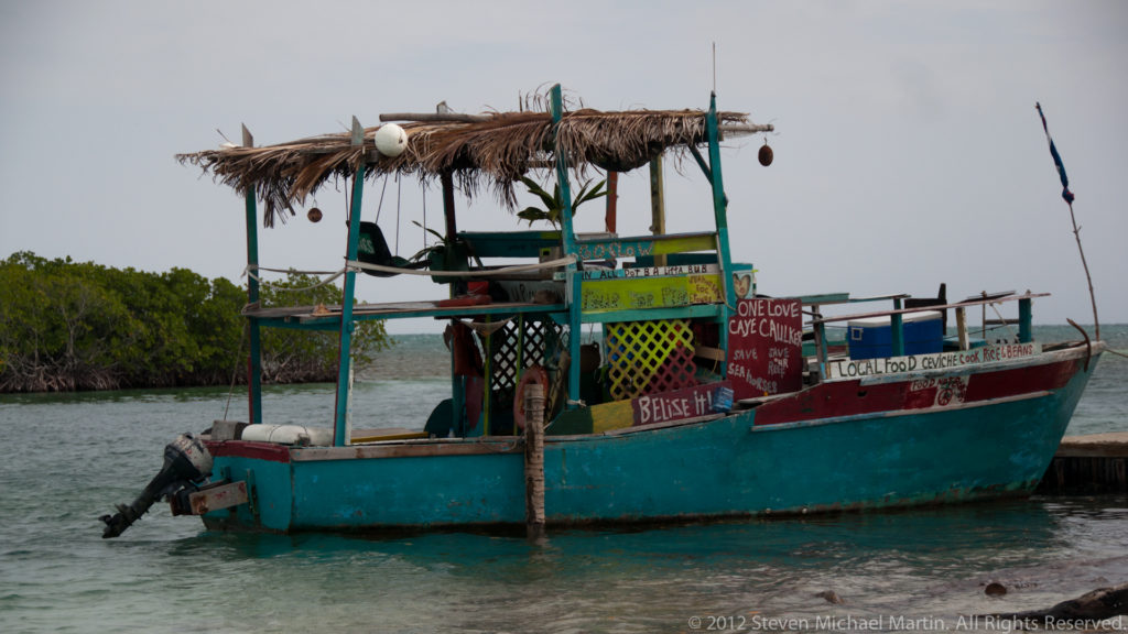 Turquoise working boat with hand-painted lettering, canopy, ropes, and outboard motor moored beside a dock.
