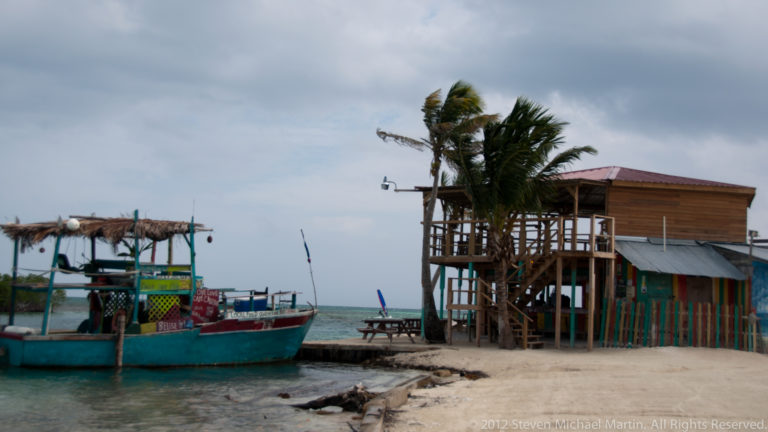 Painted boat moored by a dock beside a simple wooden waterfront structure and wind-blown palm trees.