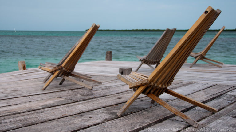 Several wooden reclining chairs arranged on a weathered dock facing turquoise Caribbean water.