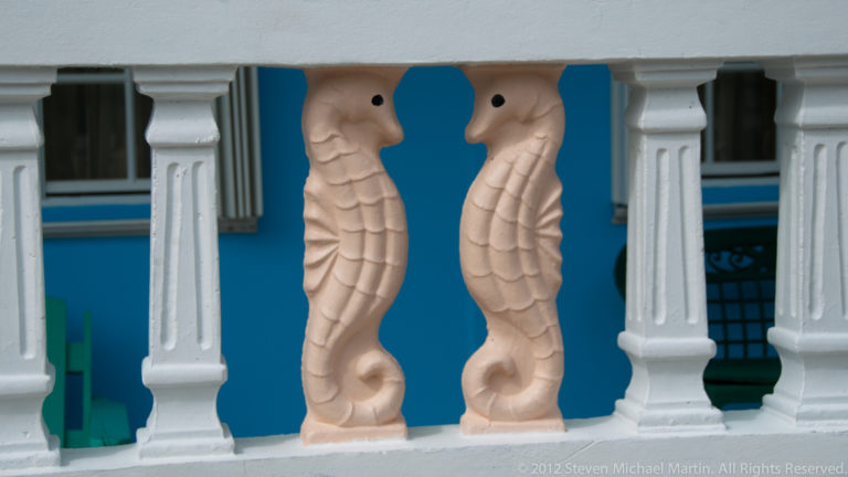 Two pale peach seahorse-shaped balusters between white railing posts against a bright blue wall.