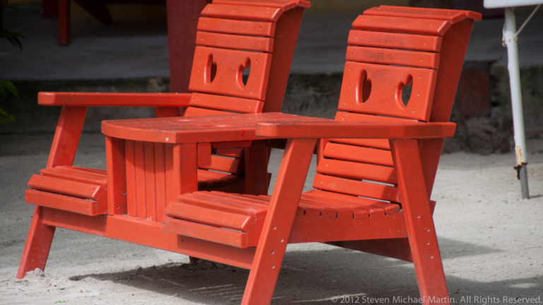 Bright red paired Adirondack-style chairs joined by a small center table on pale sandy ground.