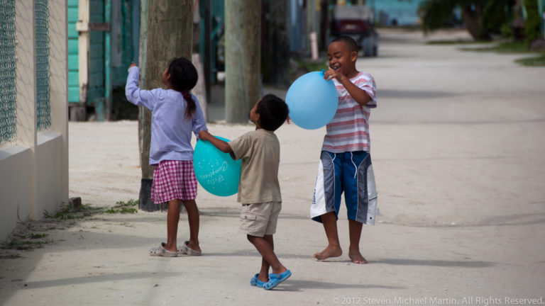 Three children playing with blue balloons on a sandy residential street in Caye Caulker.