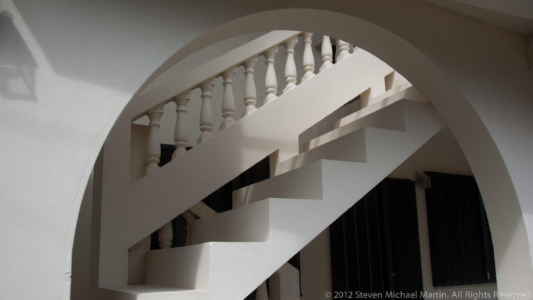 White exterior staircase crossing a large masonry arch, with turned balusters and strong sunlit shadows.