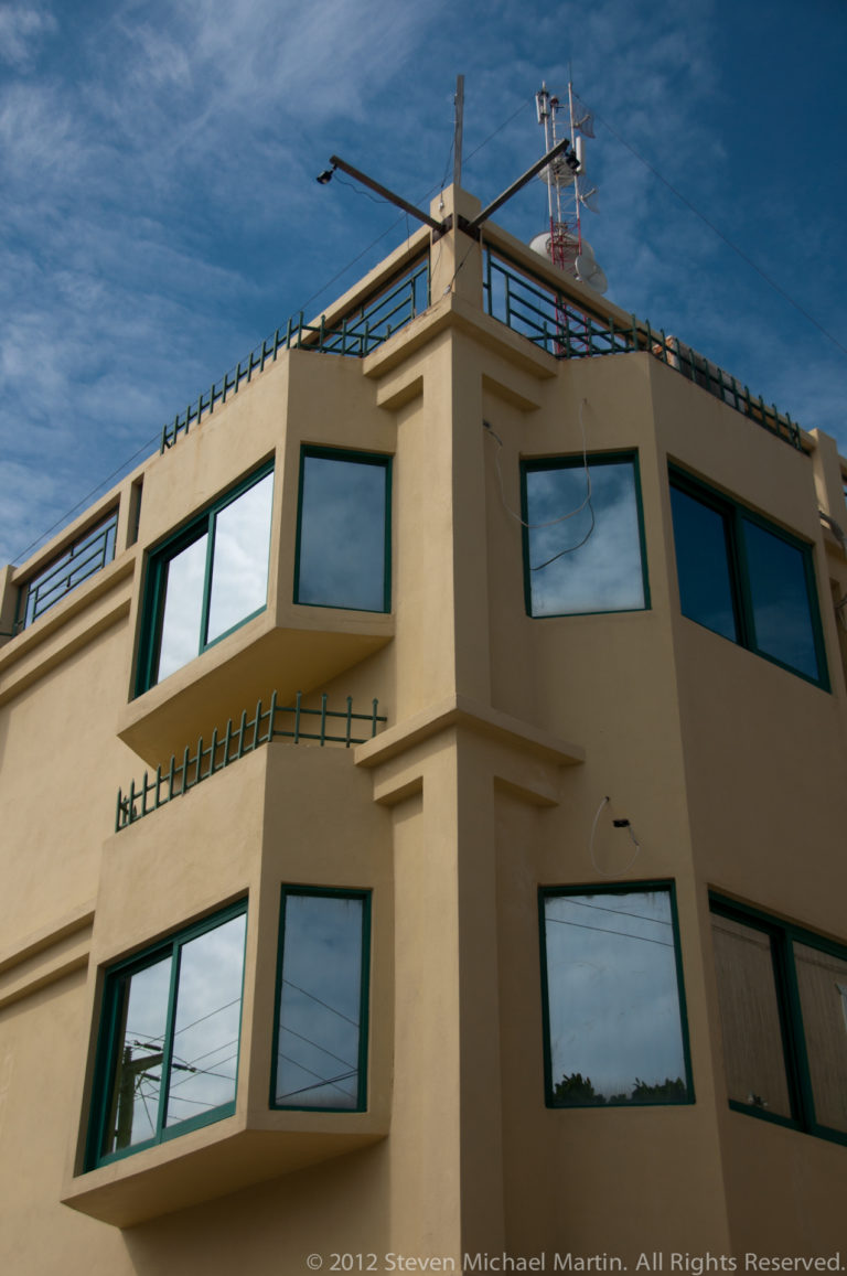Upward view of a beige multi-story building with projecting bays, green-framed windows, rooftop railings, and antennas.