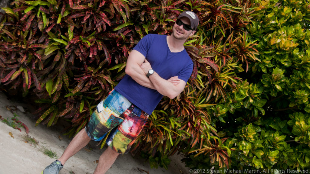 Person in blue shirt and colorful shorts leaning beside dense multicolored tropical foliage on Caye Caulker.