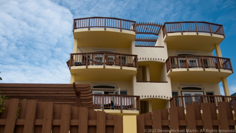 Low-angle view of a cream multi-story building with projecting balconies, rooftop terraces, and dark wooden railings behind a fence.