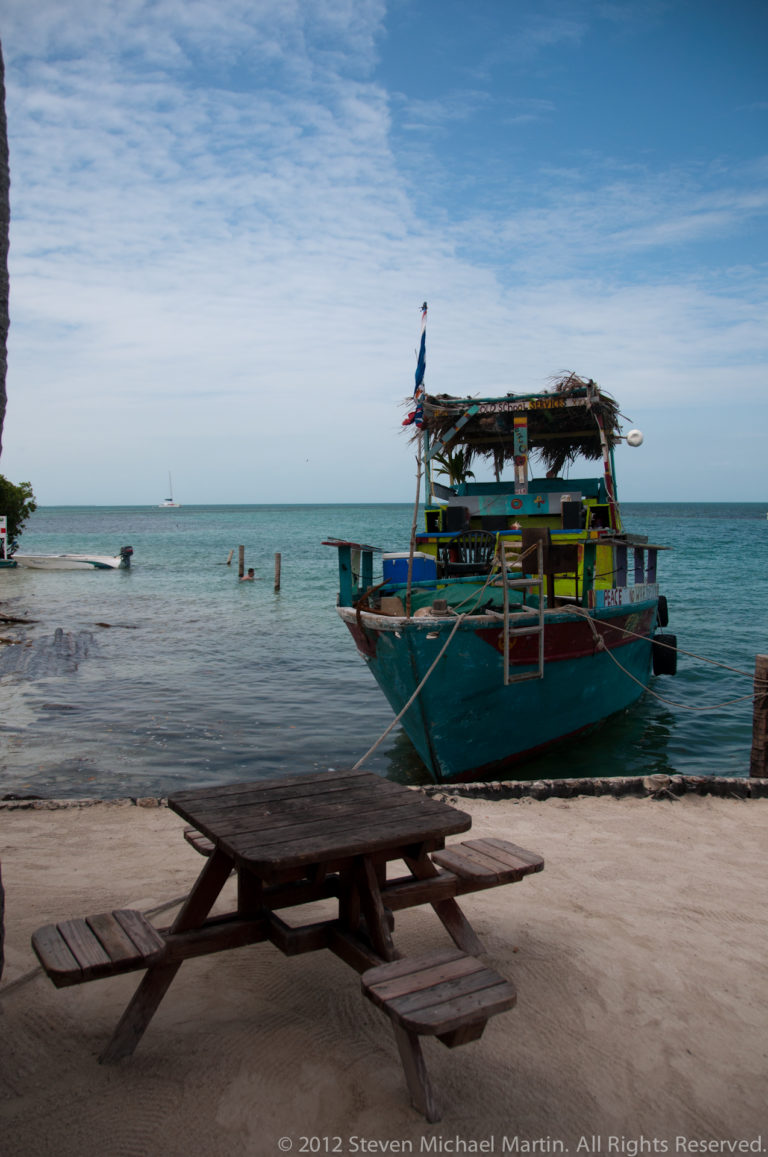 Colorful turquoise boat moored in shallow water beyond a wooden picnic table on a sandy Caye Caulker beachfront.