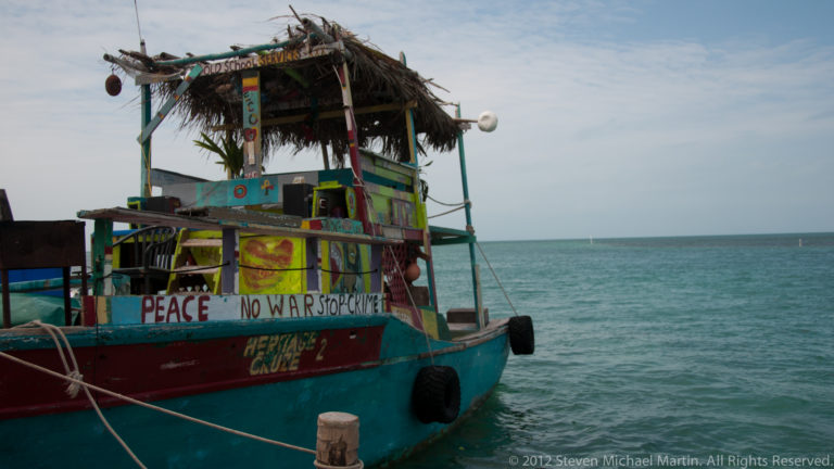 Close view of a brightly painted working boat with thatched canopy, tire fenders, ropes, and the words “PEACE” and “NO WAR.”