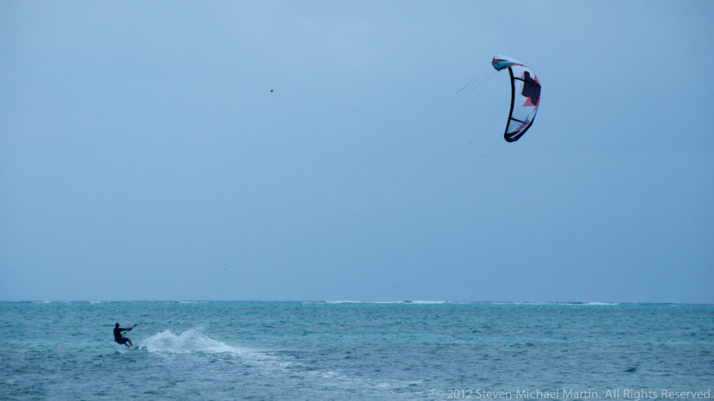 Kitesurfer moving across blue-green water beneath a large curved traction kite high in the sky.