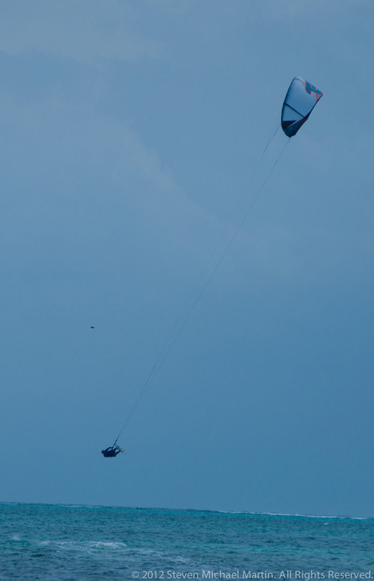 Wide view of a distant kitesurfer on turquoise water connected by long lines to a blue kite high overhead.