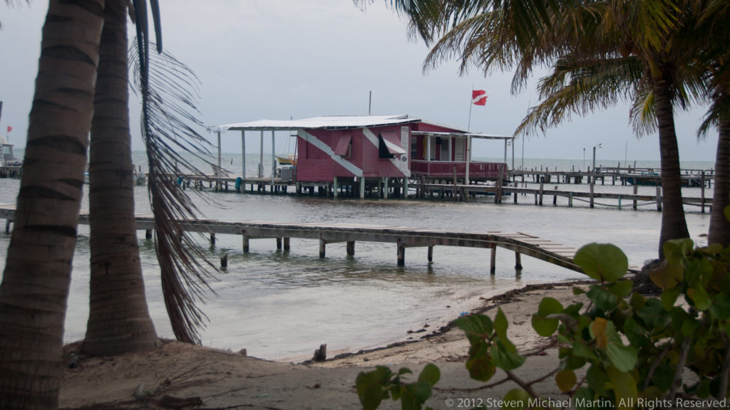 Pink wooden building on pilings over shallow water, reached by long narrow piers and framed by coconut palms.