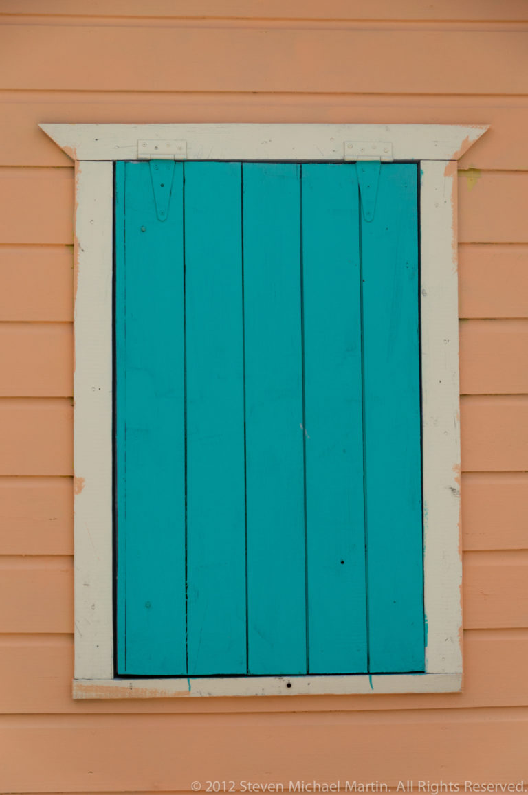 Closed turquoise plank shutters with white trim set into a peach-colored horizontal clapboard wall.