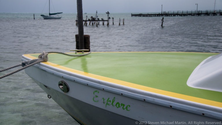 Bow of a boat named Explore with a green deck and yellow trim, moored beside posts with a pier across the water.