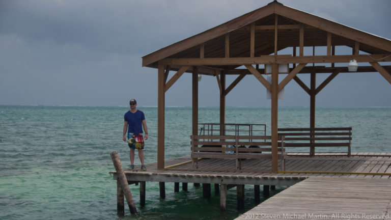 Wooden open-sided shelter and bench on a pier over turquoise water, with a person walking along the dock.