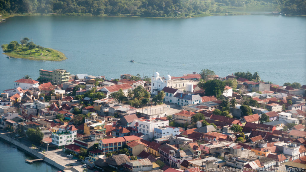 Aerial view of Flores, Guatemala, with dense red-roofed buildings beside blue Lake Petén Itzá and wooded shoreline beyond.