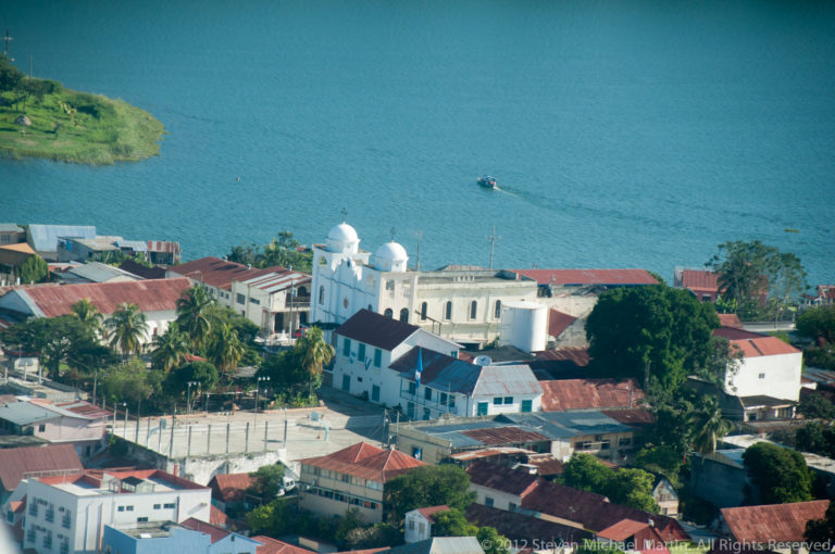 Elevated view of a white twin-domed church among Flores rooftops beside the blue water of Lake Petén Itzá.