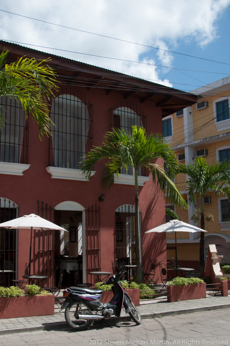 Red stucco building with barred windows, palm planters, white patio umbrellas, shaded entrance, and a motorcycle parked in front.