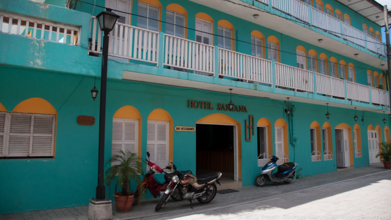 Turquoise Hotel Santana façade with ochre arches, white shutters and balcony railings, and motorcycles parked near the entrance.