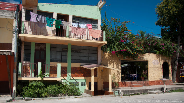 Two-story colorful residence in Flores with green columns, balcony laundry, shaded ground floor, and flowering vegetation.