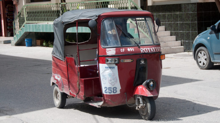 Weathered red three-wheeled tuk-tuk taxi with black canopy parked on a sunny street in Flores, Guatemala.