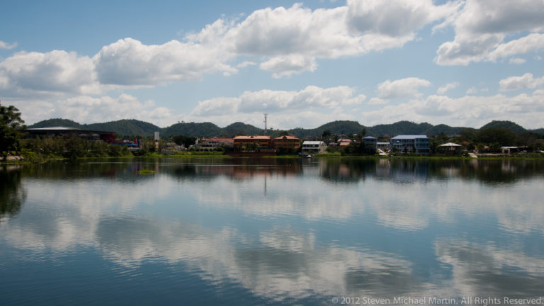 Calm Lake Petén Itzá reflecting white clouds and blue sky, with low buildings and wooded hills along the far shore.