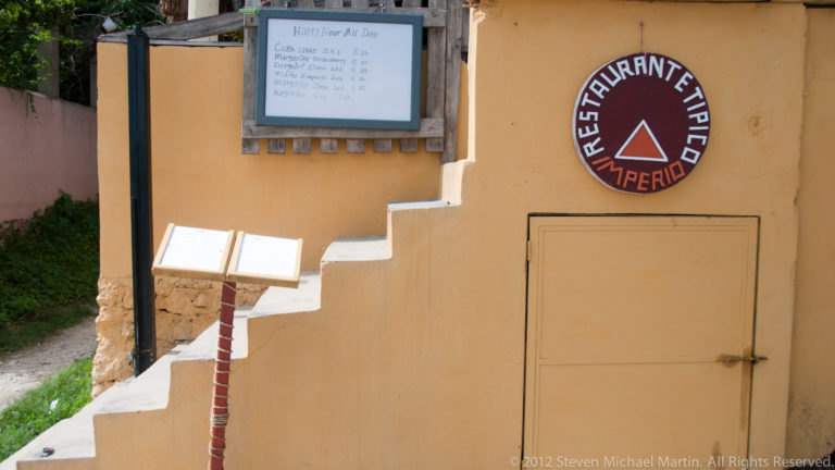 Circular Restaurante Típico Imperio sign on an ochre wall beside a small stairway and posted menu in Flores.