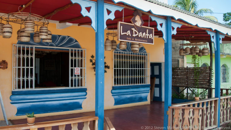 Colorful La Danta restaurant façade with ochre walls, blue trim, barred windows, wooden veranda, and hanging barrel decorations.