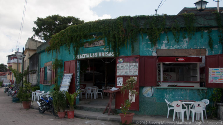 Placita Las Brisas restaurant in a weathered turquoise storefront with open windows, menu boards, chairs, plants, and motorcycles in Flores.