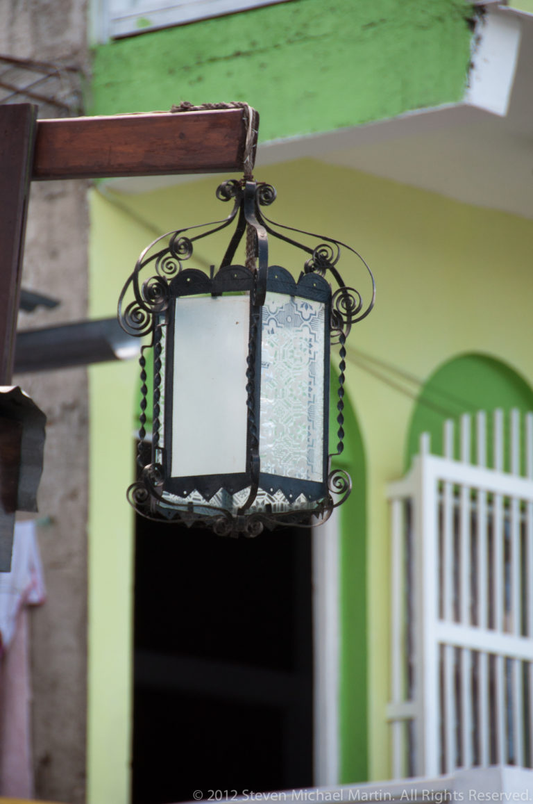 Decorative dark metal lantern with frosted glass panels hanging before a pale green building and white barred window in Flores.