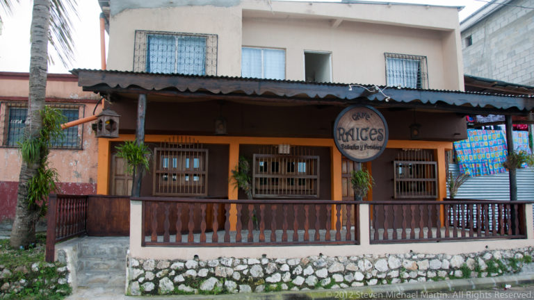 Façade of Raíces restaurant in Flores with wooden veranda, oval sign, dark timber details, potted plants, and stone base.
