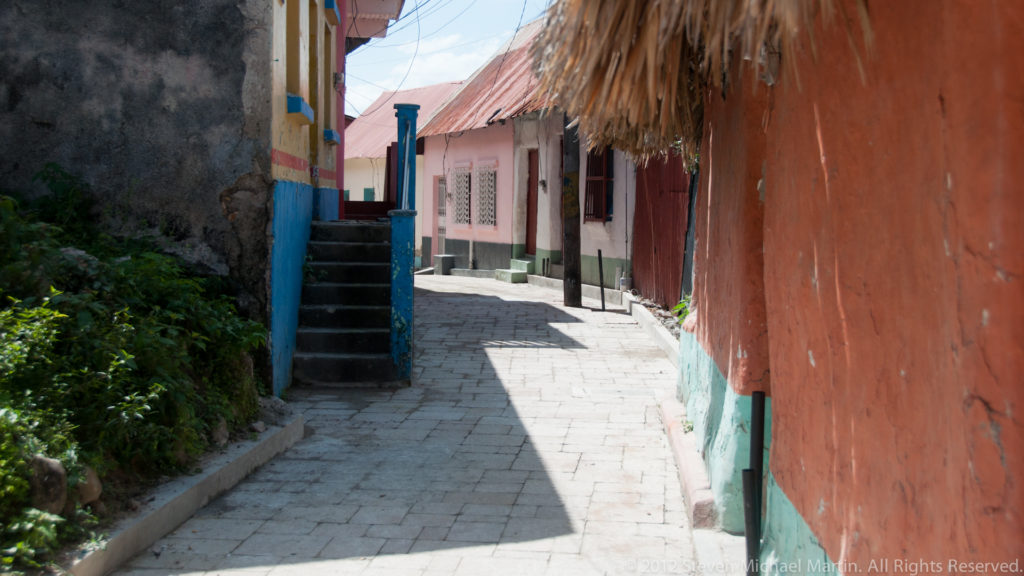 Narrow paved lane between orange, turquoise, pink, and white buildings in Flores, Guatemala, with small steps and vegetation along one side.