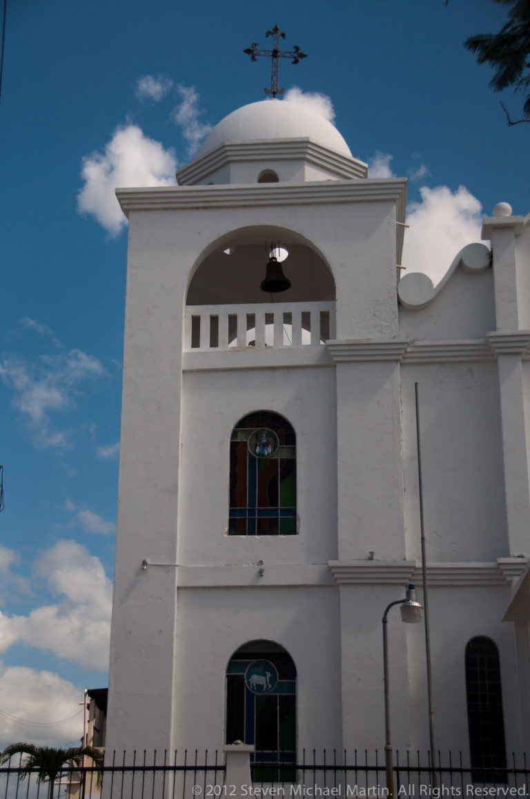 Frontal white church façade with bell arch, stained-glass windows, dome, and metal cross beneath a blue sky in Flores.