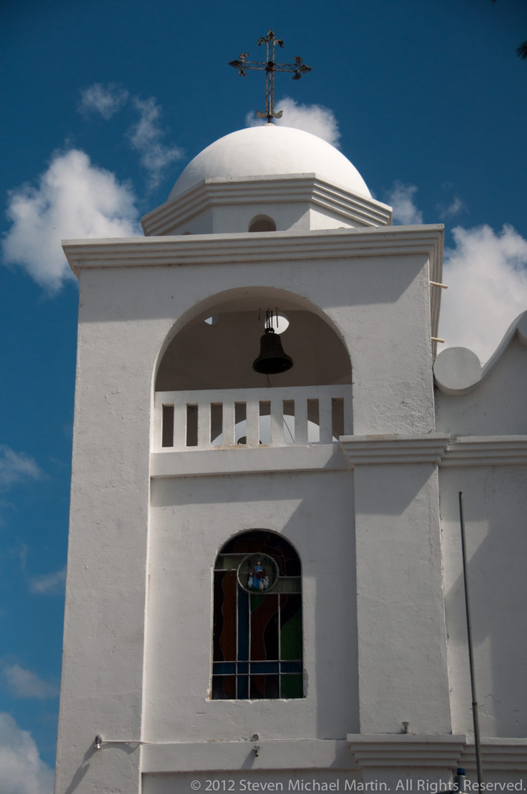 Upper white church façade with rounded dome, metal cross, open bell arch, and stained-glass window in Flores, Guatemala.
