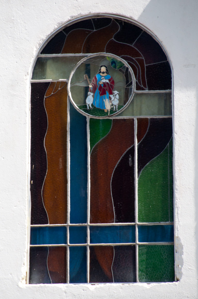 Arched stained-glass church window with multicolored panes and a small central devotional figure set in a white stucco wall.