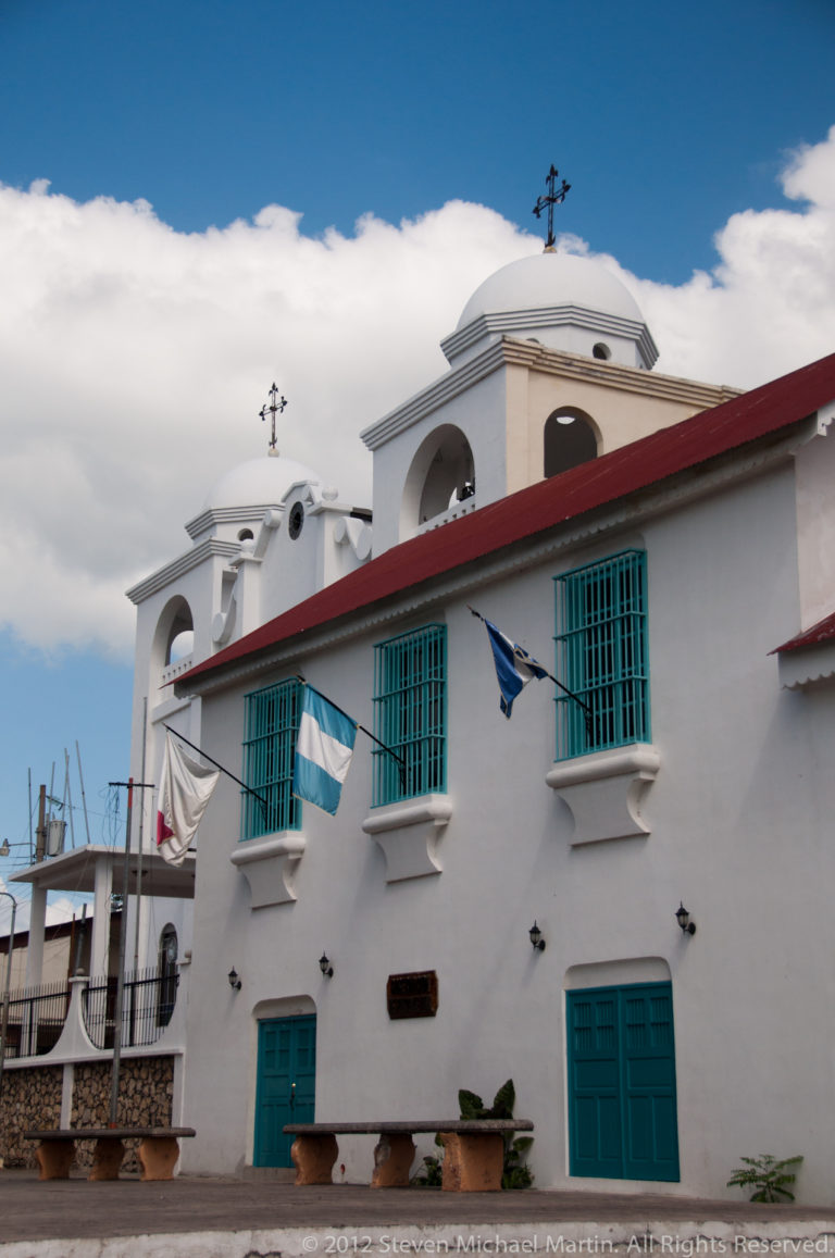 White stucco church façade in Flores with blue-green shutters, curved parapets, arched upper opening, and Guatemalan flags.