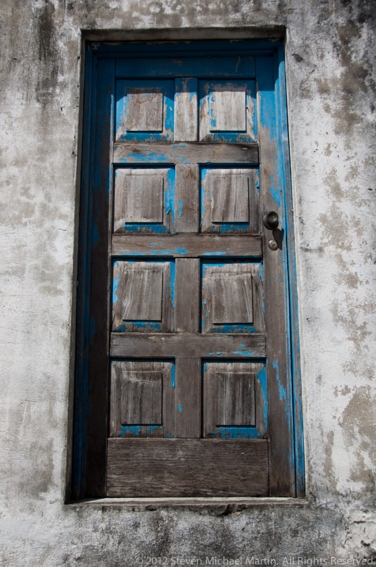 Weathered paneled wooden door with peeling blue paint set into a rough pale wall in Flores, Guatemala.
