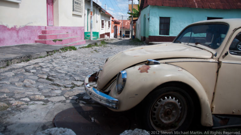 Weathered Volkswagen Beetle beside a narrow cobblestone street lined with brightly painted houses in Flores, Guatemala.