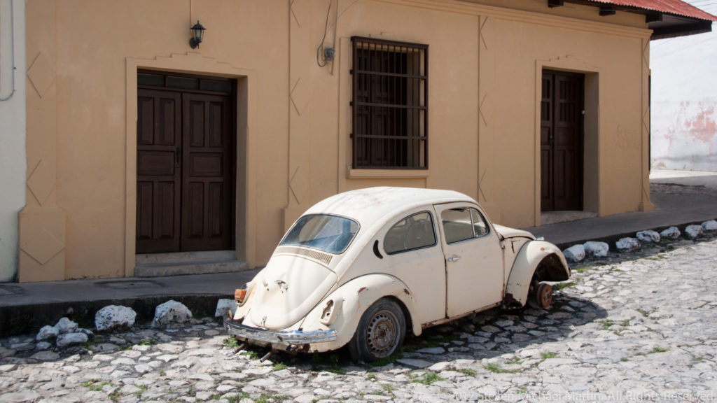 Weathered white Volkswagen Beetle parked on a cobblestone street beside a pale stucco building with dark wooden doors in Flores.