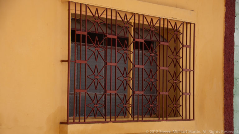 Decorative reddish-brown iron window grille with star and diamond patterns set into an ochre wall in Flores, Guatemala.