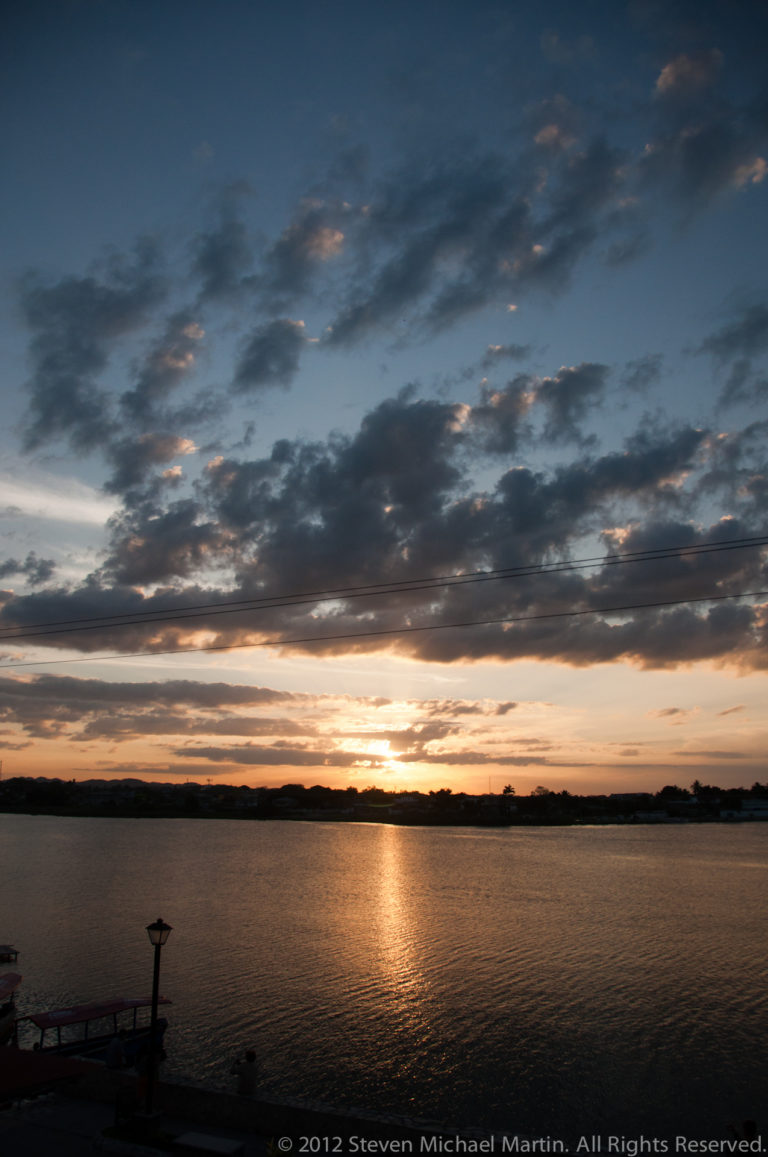 Lake Petén Itzá at sunset with a soft orange reflection, broken clouds, and a small distant boat near the horizon.