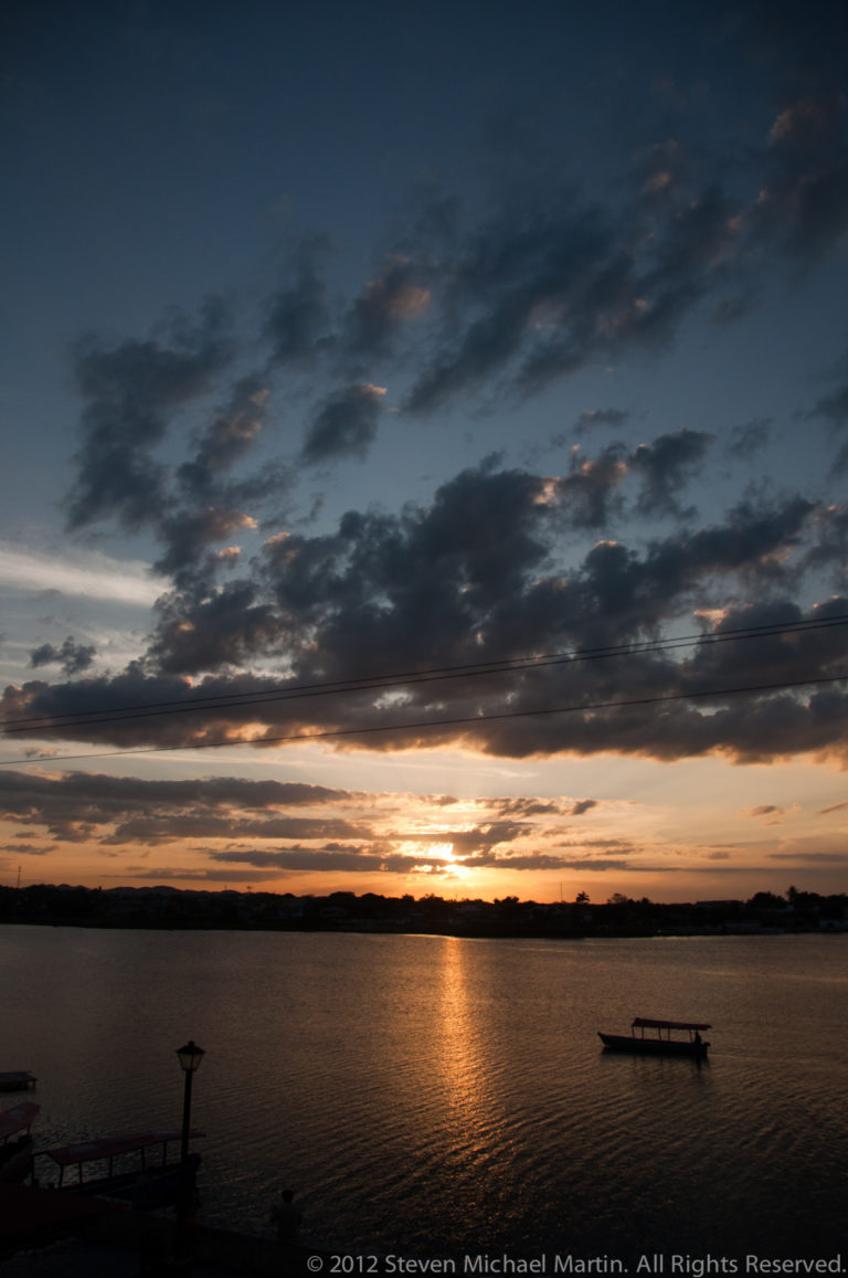 Wide sunset view across Lake Petén Itzá with layered clouds, a long golden reflection, and small boats near the horizon.