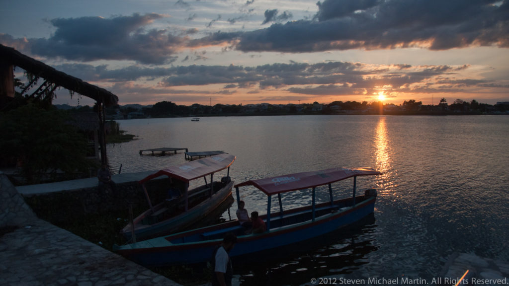 Covered passenger boats moored at the Flores waterfront during sunset, with a bright reflection extending across Lake Petén Itzá.