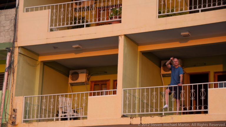 Man standing on a balcony of a pale stucco building in Flores, with white railings, doors, and air-conditioning units.