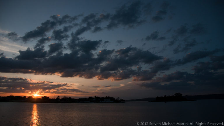 Setting sun beneath dark clouds casts a narrow golden reflection across Lake Petén Itzá toward the camera.
