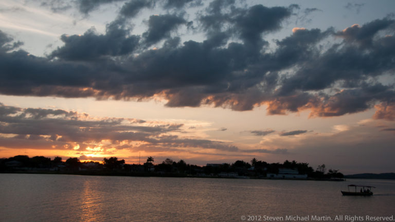 Small boat crossing Lake Petén Itzá under dark clouds with warm sunset light near the far shore.