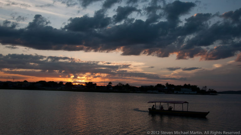 Covered passenger boat silhouetted on Lake Petén Itzá beneath dark clouds and a glowing sunset horizon near Flores, Guatemala.