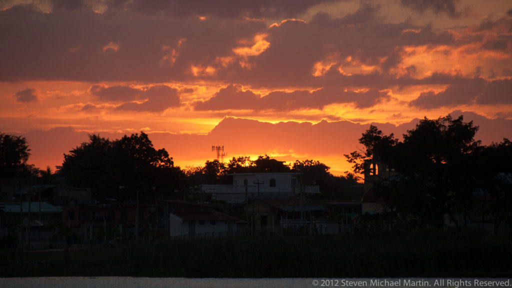Vivid orange sunset behind dark rooftops and trees in Flores, with glowing clouds and a small communications structure in silhouette.