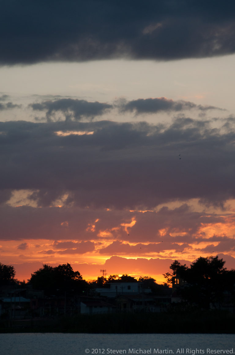 Orange sunset beneath dark layered clouds over a silhouetted lakeshore with low buildings and trees in Flores, Guatemala.