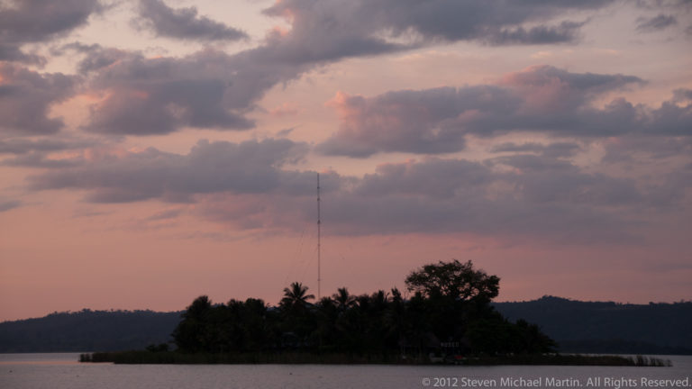 Pink and lavender clouds at dusk above Lake Petén Itzá, with a dark shoreline, palms, and a slender communications mast.